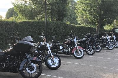 Plusieurs motos stationnées dans un parking entouré d'arbres, avec un véhicule utilitaire en arrière-plan.