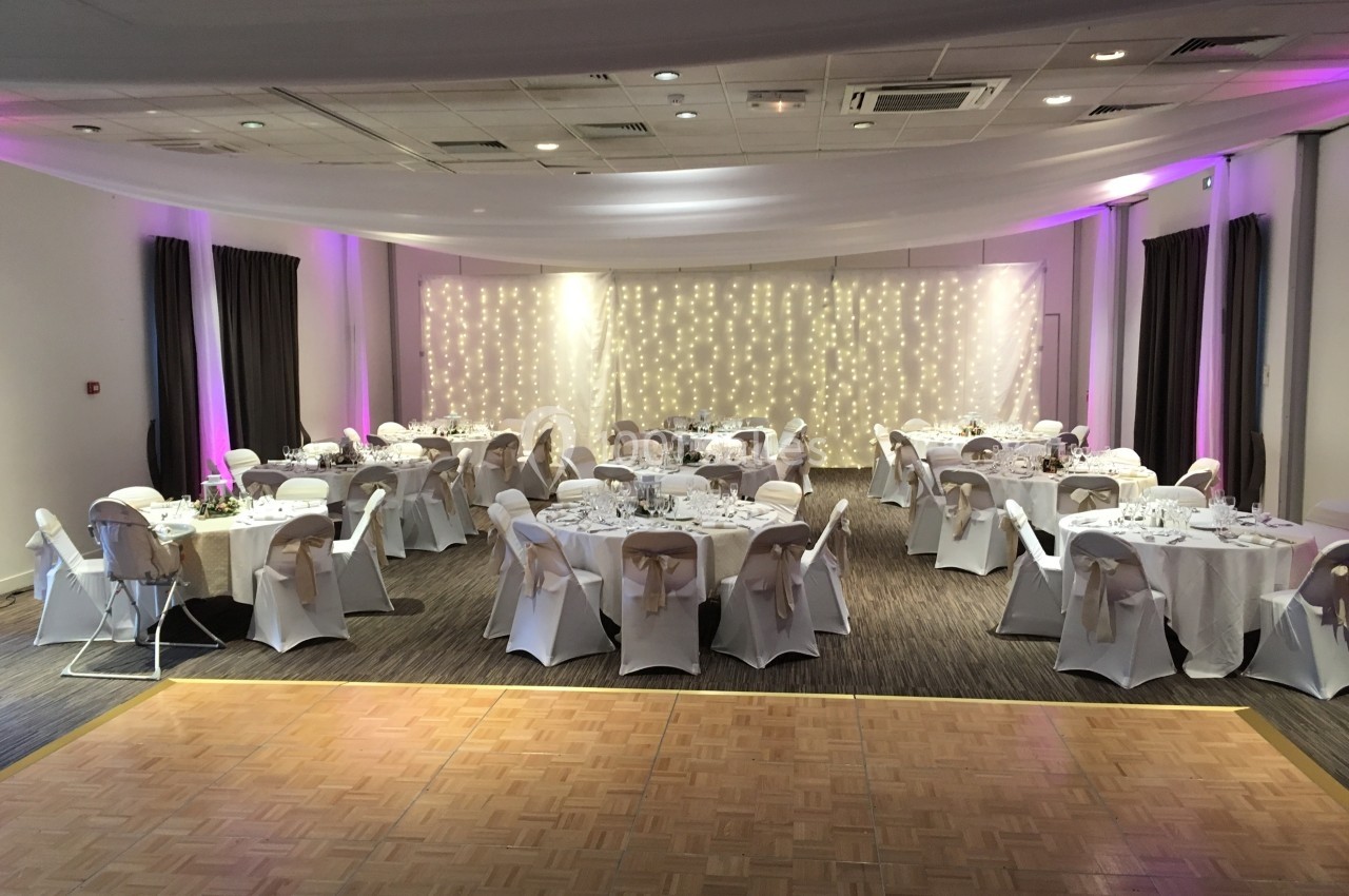 Location salle Saint-Martin-du-Vivier (Seine-Maritime) - The Originals Relais La Bertelière - Rouen #30 Salle de réception décorée avec des tables rondes dressées et un mur illuminé par des guirlandes lumineuses.