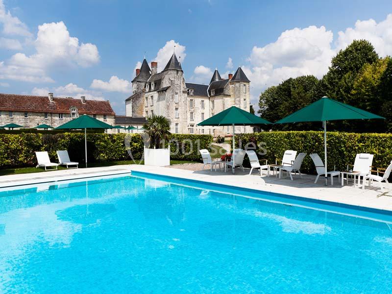 Piscine extérieure entourée de chaises longues, parasols verts et vue sur un château en pierre sous un ciel bleu.