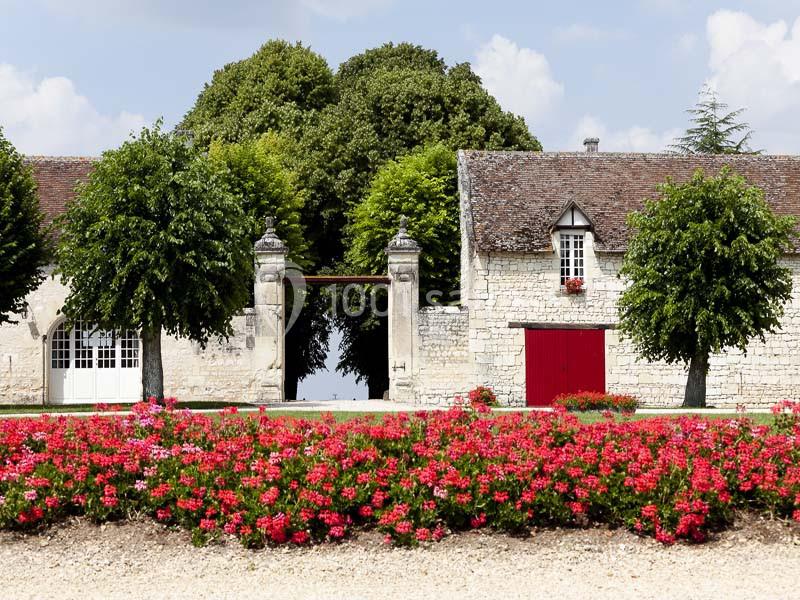 Bâtiment en pierre avec toit en tuiles, entouré d'arbres et d'un parterre de fleurs rouges sous un ciel dégagé.