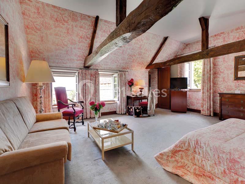 Chambre spacieuse avec poutres apparentes, canapé, lit, bureau, télévision et fenêtres donnant sur un extérieur verdoyant.