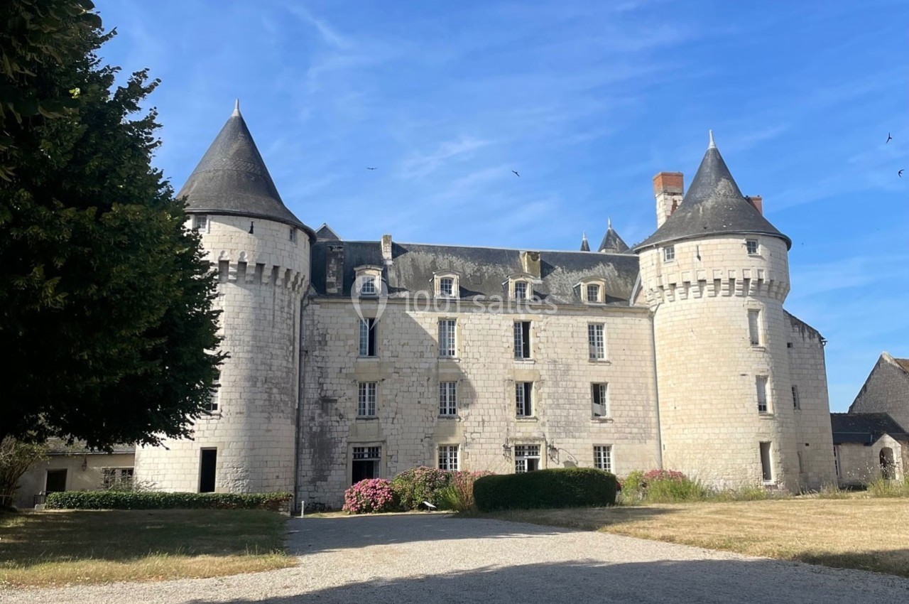 Façade d'un château en pierre blanche avec tours rondes et toit d'ardoise, entouré d'un jardin sous un ciel bleu.