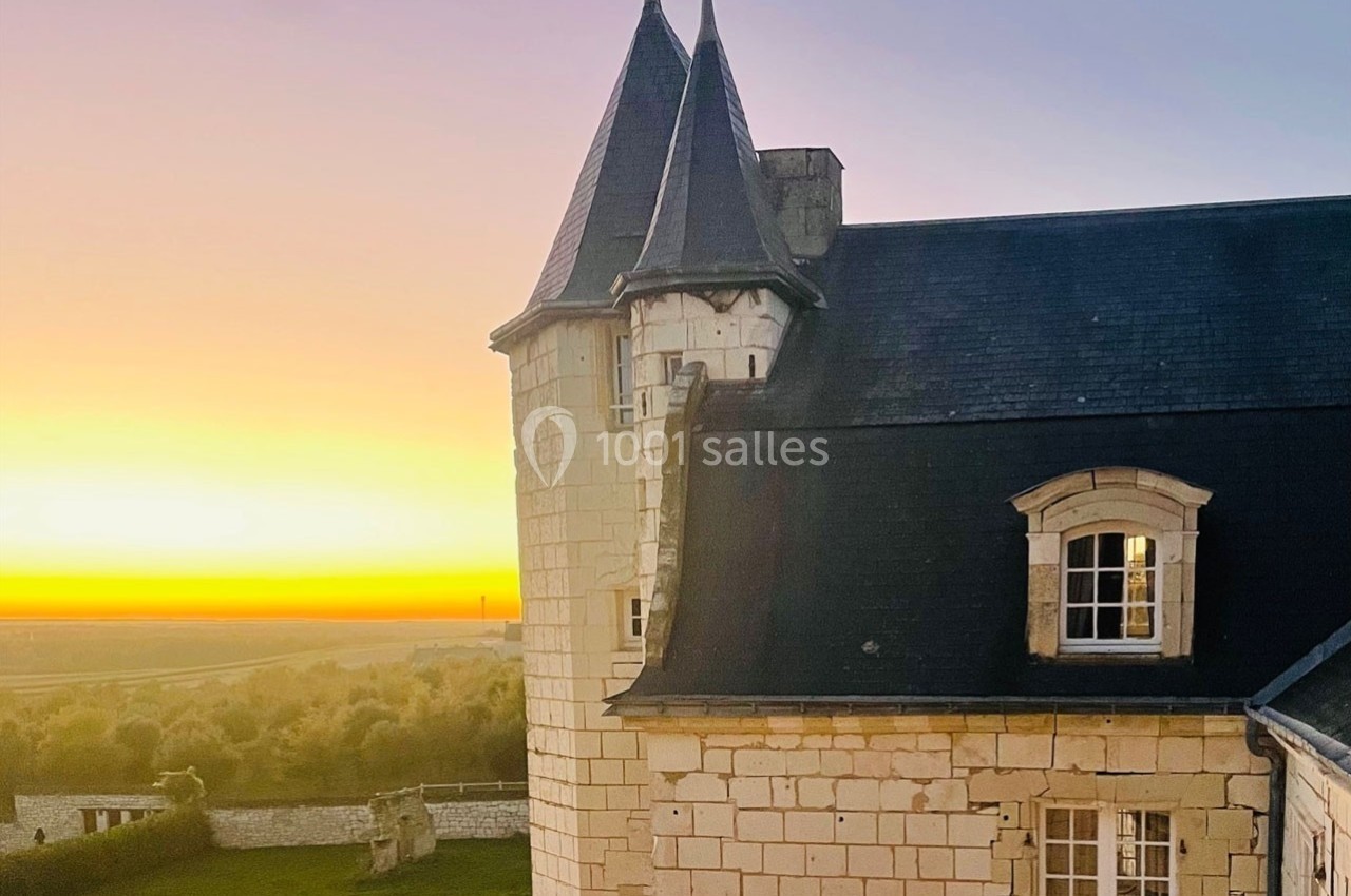Tour en pierre d'un château avec toit en ardoise, éclairée par un coucher de soleil sur un paysage rural.