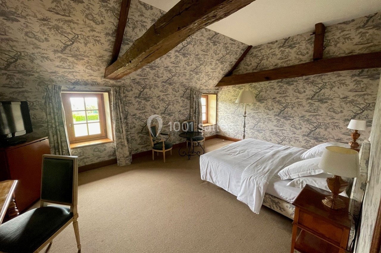 Chambre avec poutres apparentes, lit double, mobilier classique et fenêtres donnant sur un extérieur verdoyant.