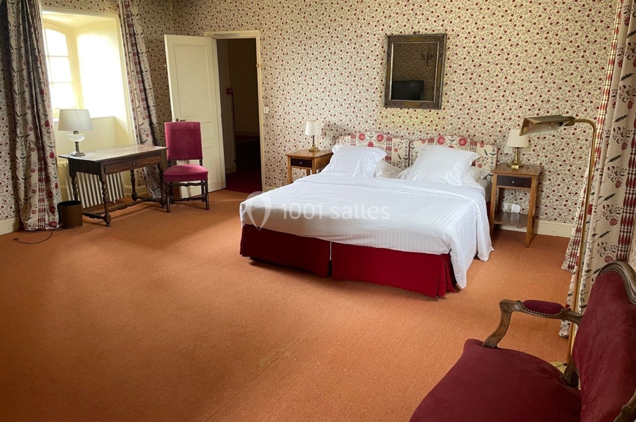 Chambre avec lit double, murs tapissés de motifs floraux, mobilier en bois et fauteuils rouges sur moquette orange.