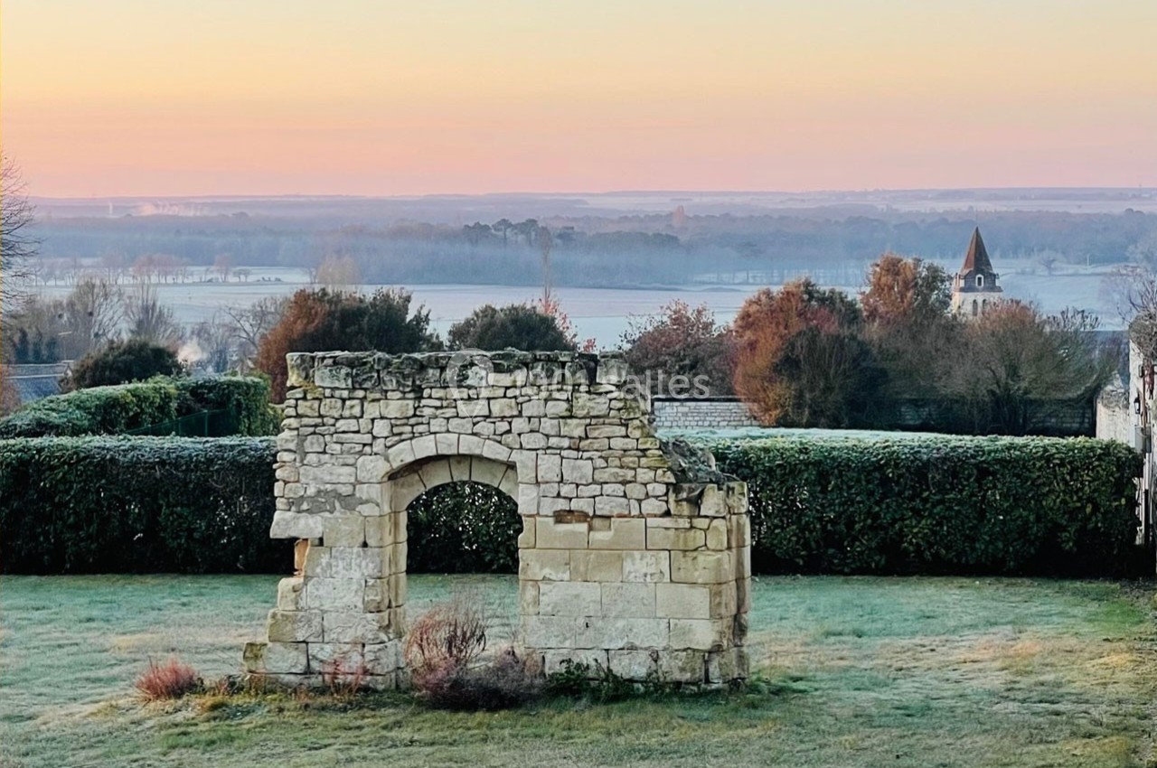 Ruine en pierre dans un jardin au lever du soleil, avec une vue dégagée sur la campagne et un clocher à l'horizon.