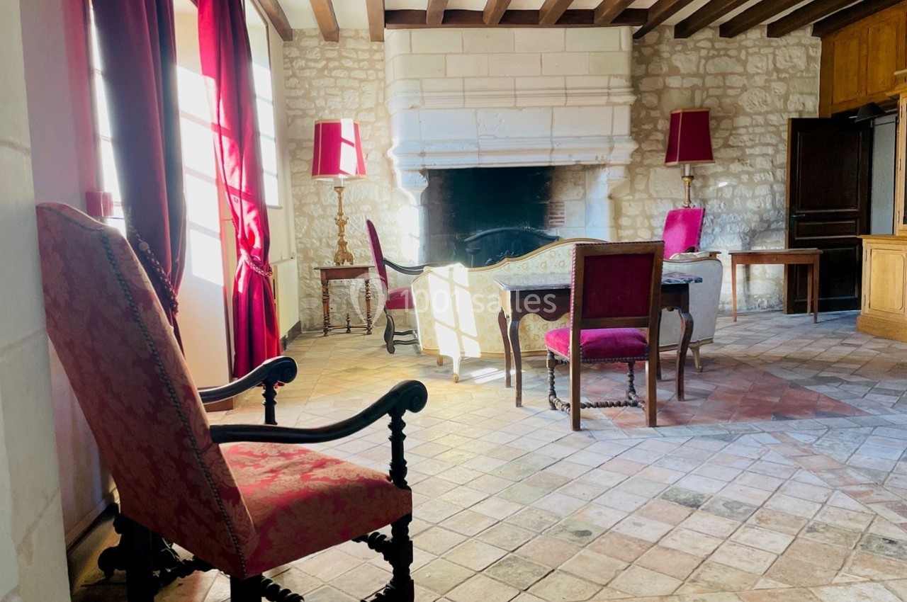 Salon lumineux avec cheminée en pierre, fauteuils et chaises en tissu rouge, sol en carreaux de terre cuite.
