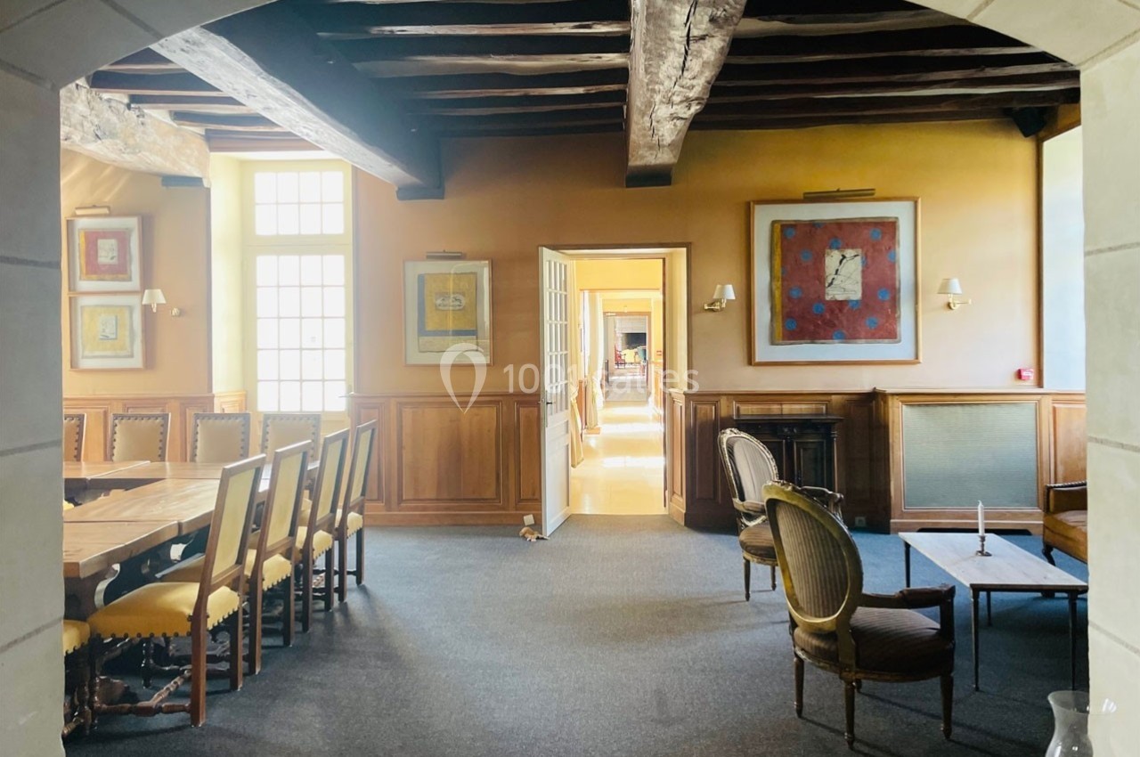 Salle lumineuse avec poutres apparentes, grande table en bois, chaises rembourrées et fauteuils près d'une cheminée.