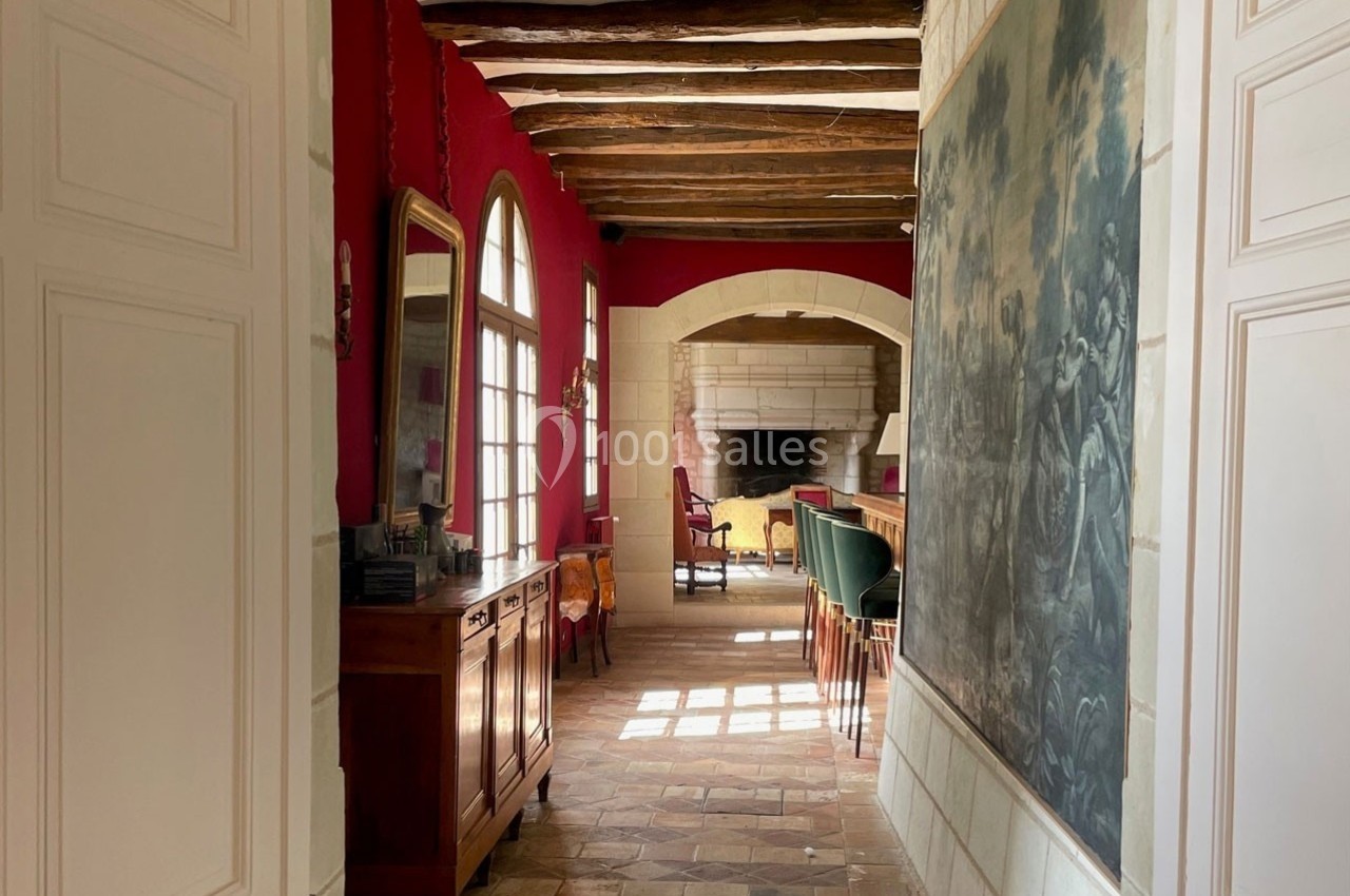Couloir avec murs rouges, poutres apparentes, meuble en bois, grande fresque murale et vue sur un salon avec cheminée.