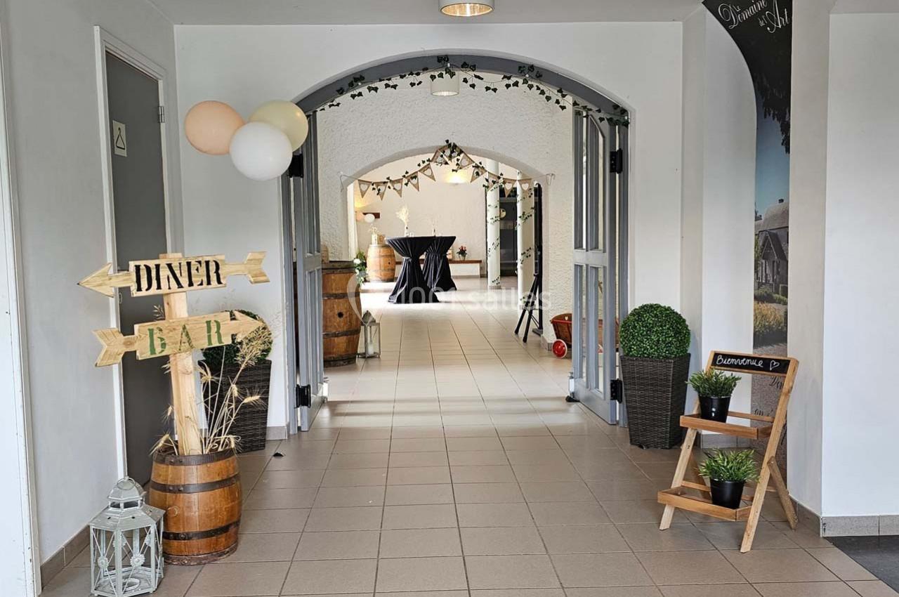 Entrée décorée avec des ballons, panneaux en bois ’Dîner’ et ’Bar’, plantes et tonneaux dans un espace lumineux.