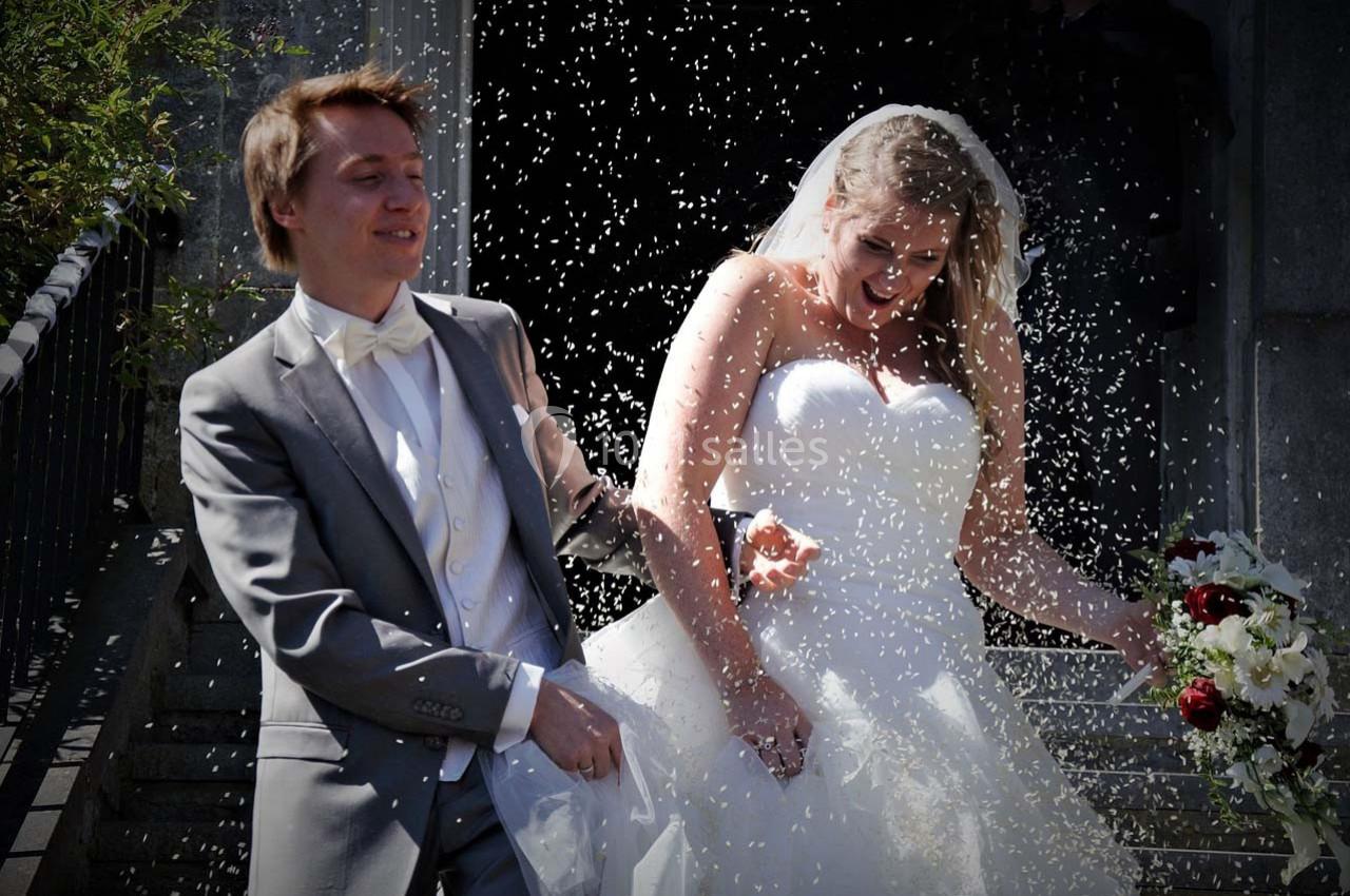 Un couple de mariés sourit en sortant d'un bâtiment sous une pluie de confettis, la mariée tenant un bouquet.