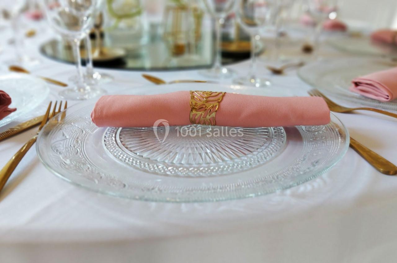Serviette rose pliée avec un anneau doré posée sur une assiette en verre transparent, entourée de couverts dorés.