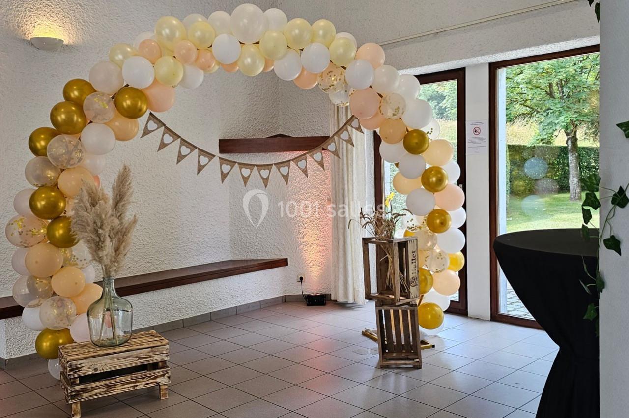 Arche décorative en ballons blancs et dorés avec des éléments en bois et des herbes séchées dans une salle lumineuse.