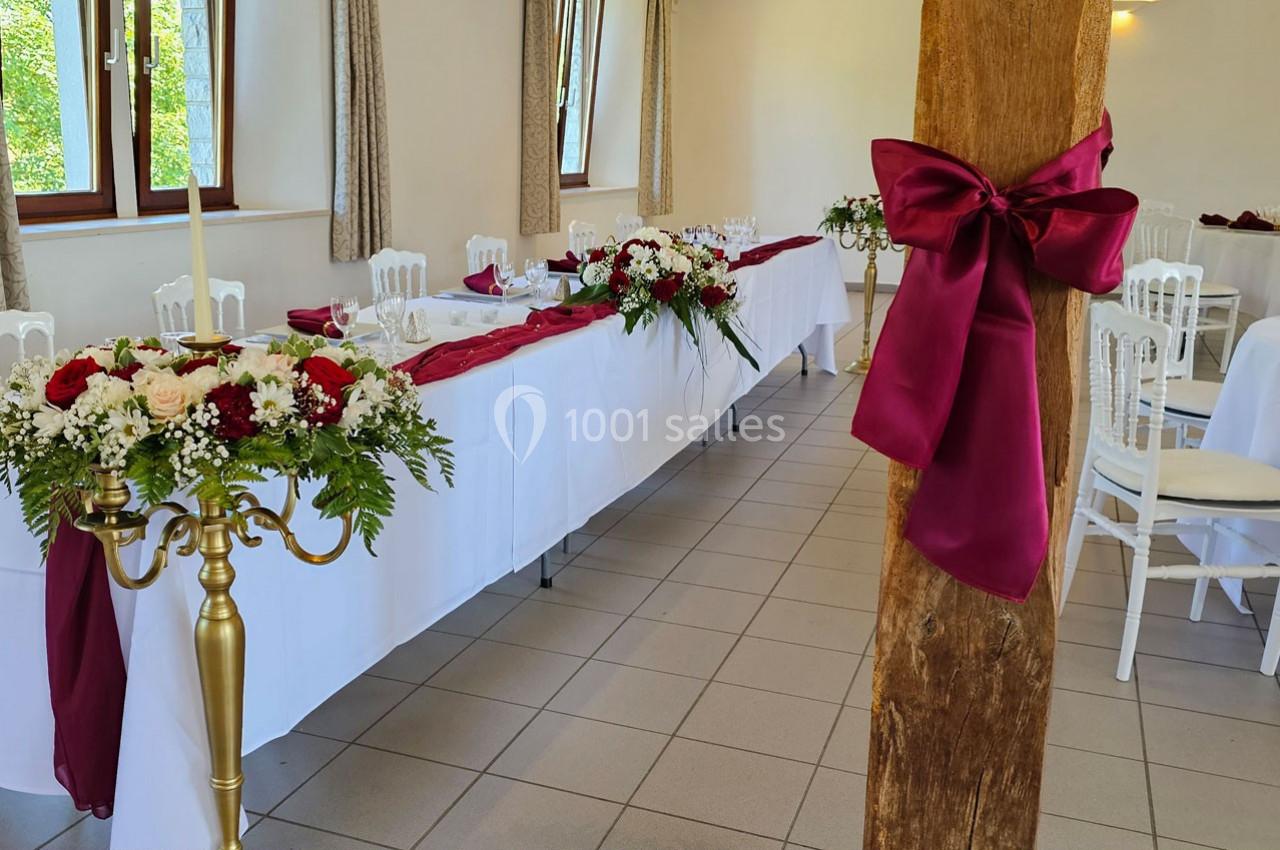 Salle de réception décorée avec des nappes blanches, des fleurs rouges et blanches, et un nœud bordeaux sur une poutre.
