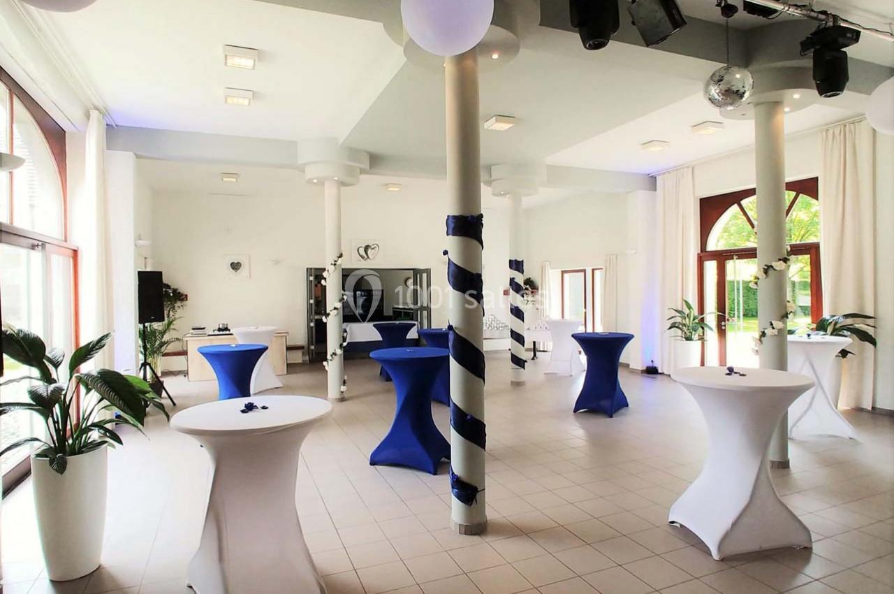 Salle lumineuse avec tables hautes drapées de bleu et blanc, décorée pour un événement, avec grandes fenêtres et plantes.