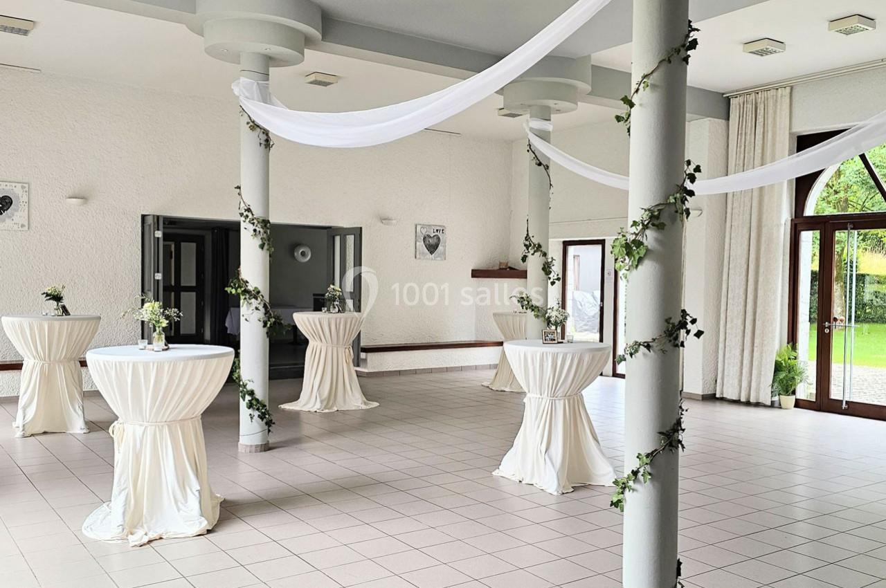 Salle lumineuse décorée de colonnes ornées de lierre et de tables hautes drapées de blanc, avec vue sur un jardin.