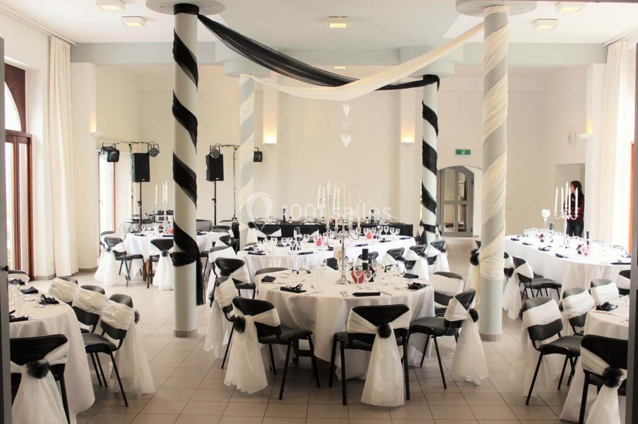 Salle de réception décorée en noir et blanc avec tables rondes, chaises habillées et colonnes ornées de rubans torsadés.