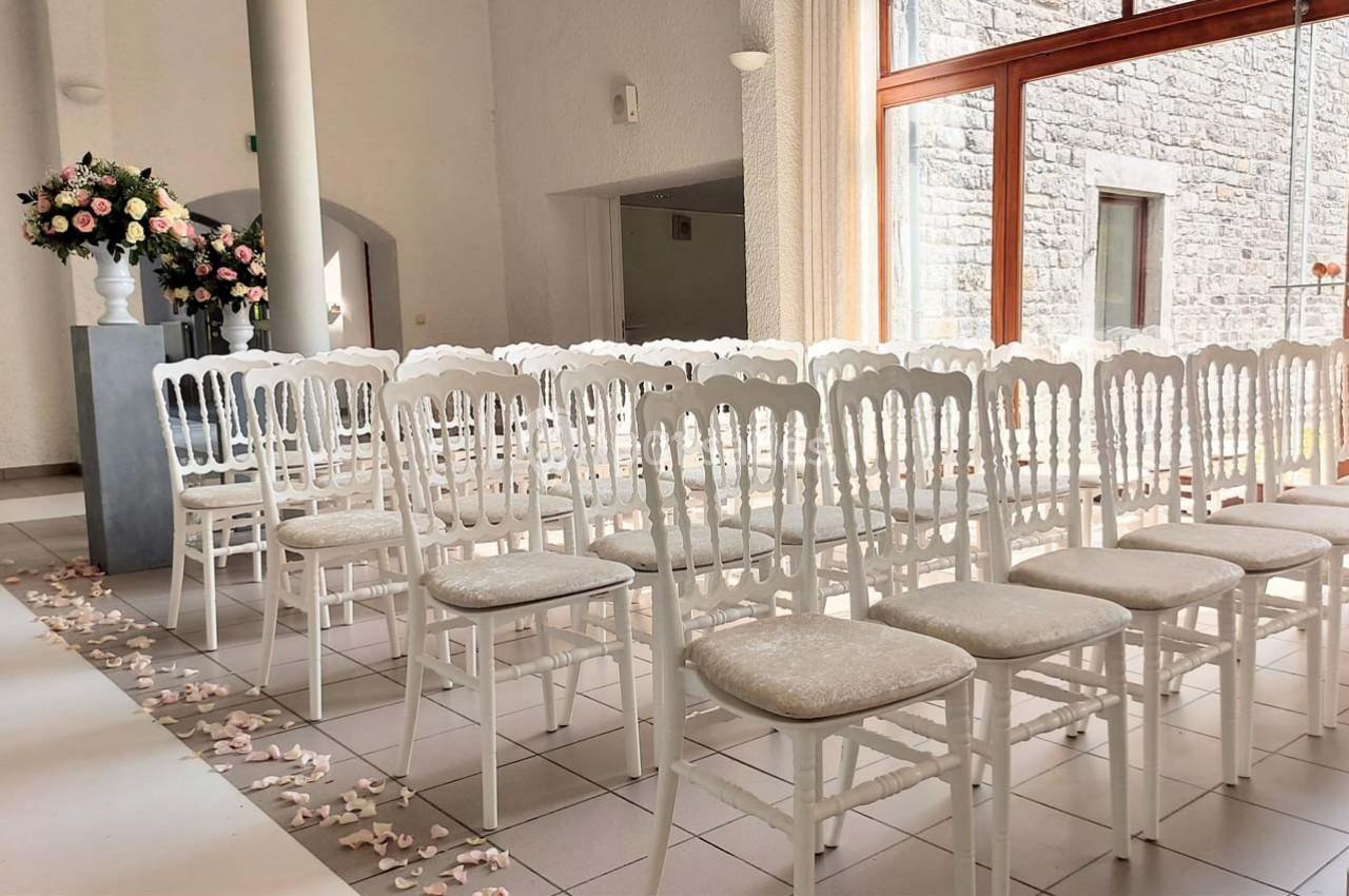 Rangées de chaises blanches alignées dans une salle lumineuse avec des pétales au sol et un bouquet floral en arrière-plan.