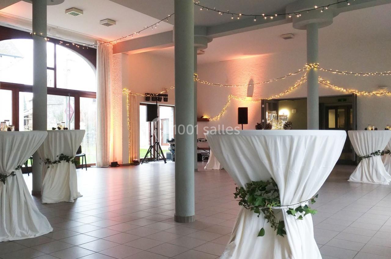 Salle de réception décorée avec des tables hautes drapées de blanc, guirlandes lumineuses et grandes baies vitrées.