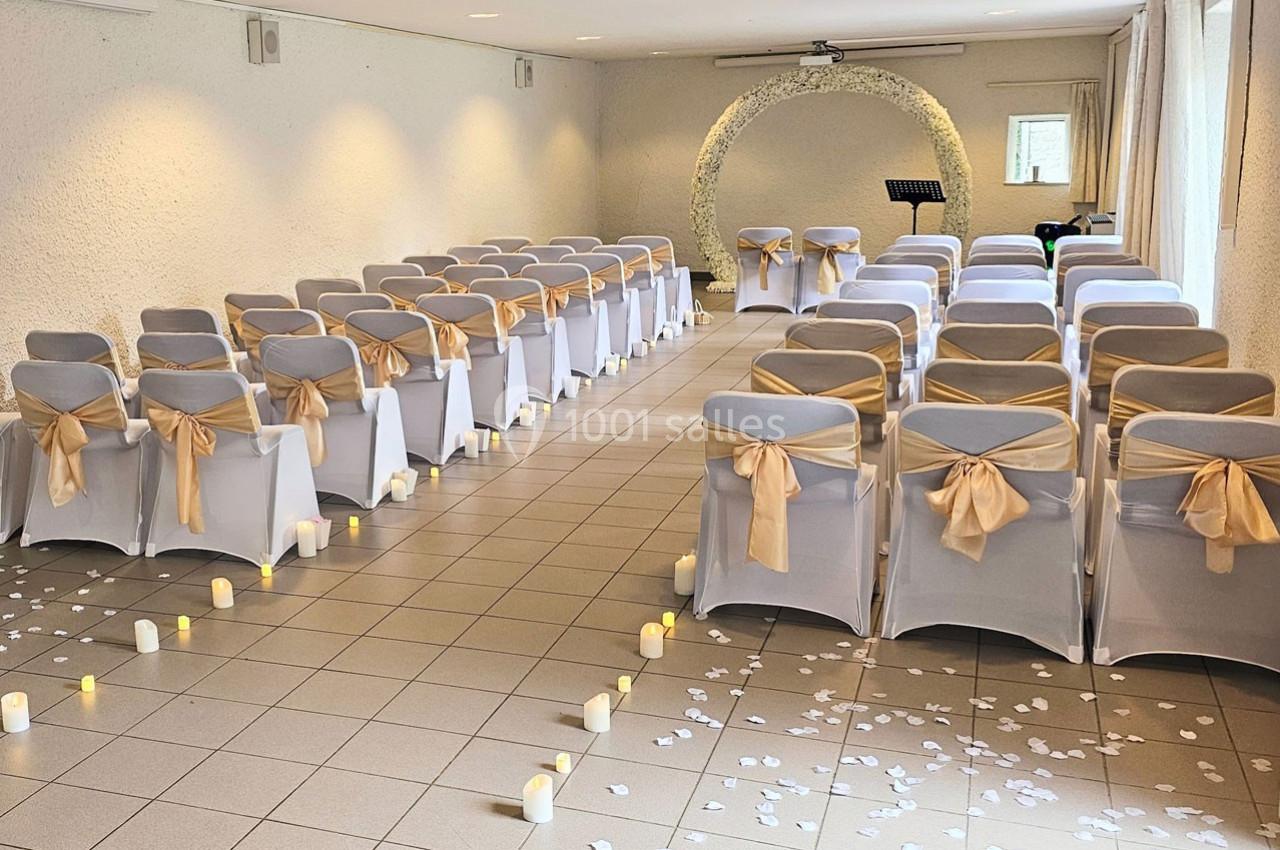 Salle décorée pour une cérémonie, avec des chaises blanches ornées de nœuds dorés, pétales au sol et bougies LED.