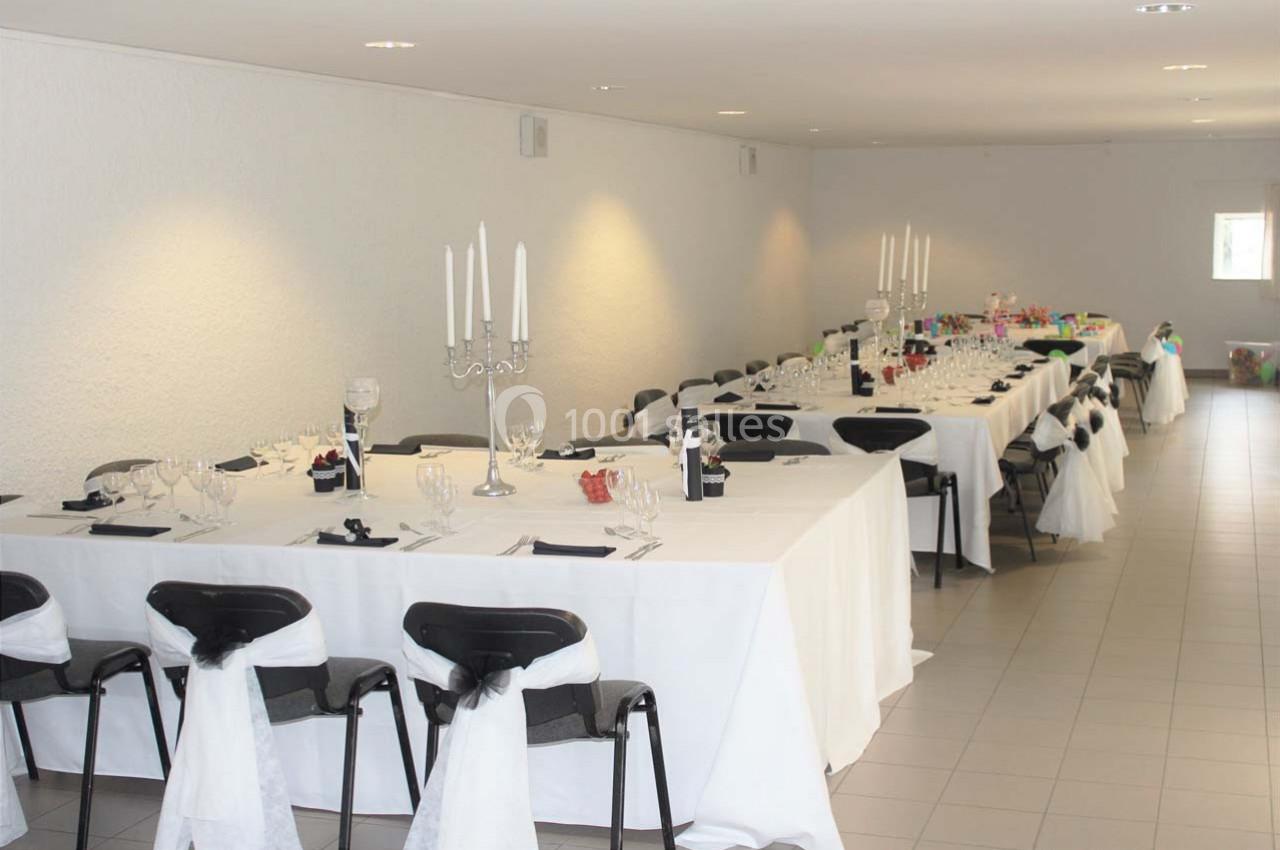 Salle décorée avec des tables alignées, nappes blanches, chaises ornées de rubans et chandeliers au centre.