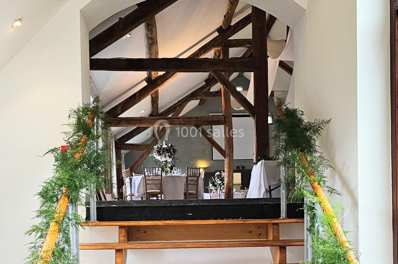 Escalier en bois décoré de verdure menant à une salle de réception avec poutres apparentes et tables dressées.
