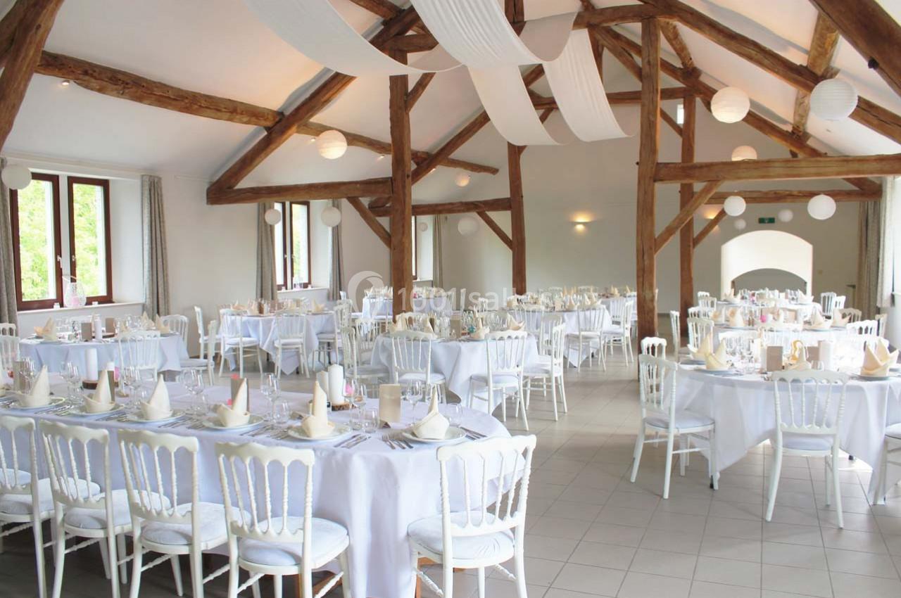 Salle de réception lumineuse avec des tables rondes dressées, chaises blanches et poutres apparentes en bois.
