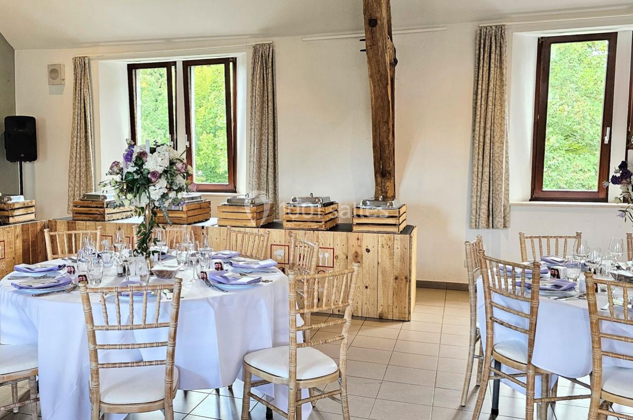 Salle de réception lumineuse avec tables rondes dressées, chaises en bois clair et décorations florales élégantes.