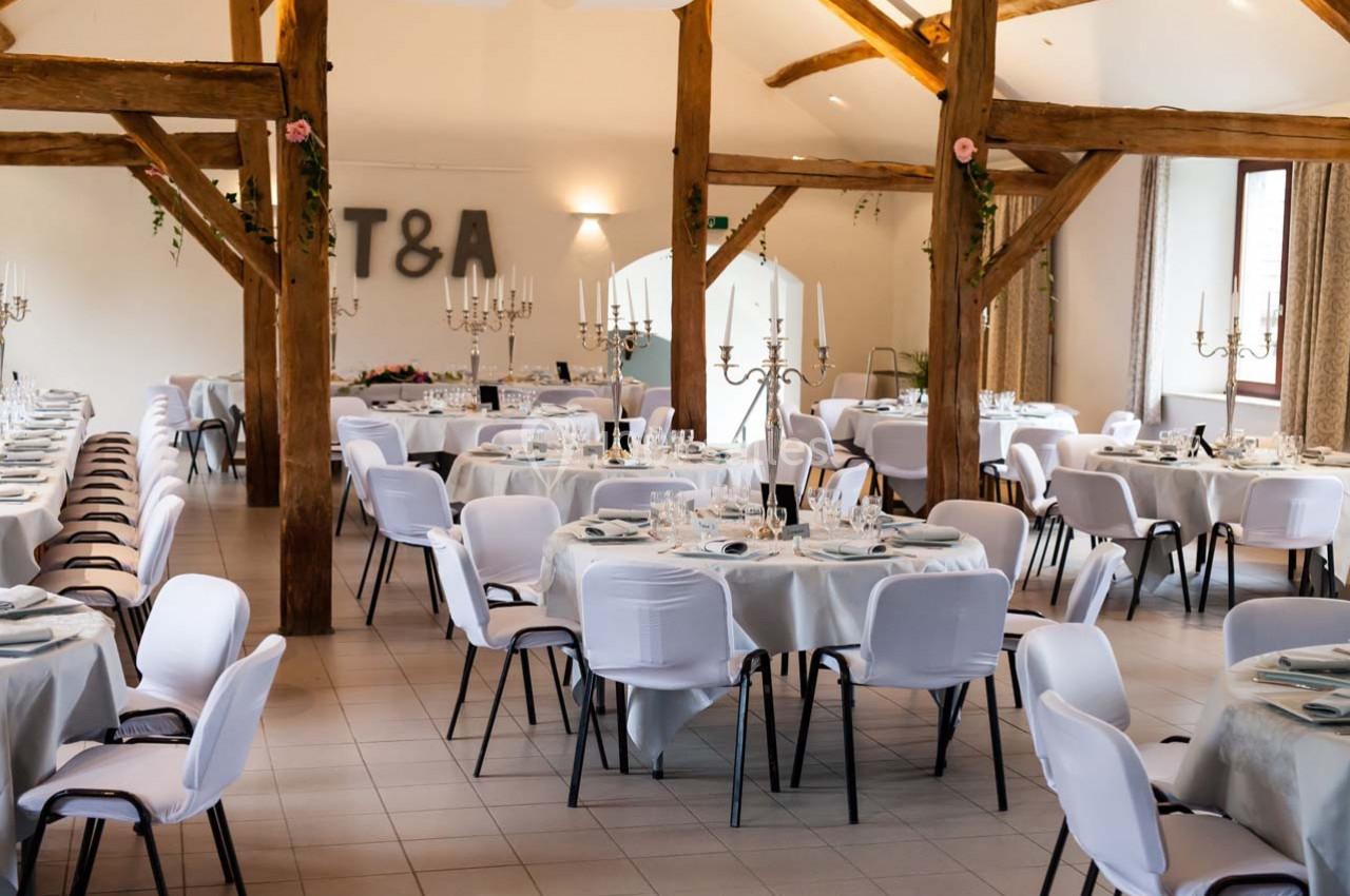 Salle de réception décorée avec des tables rondes dressées, chaises blanches et poutres apparentes en bois.
