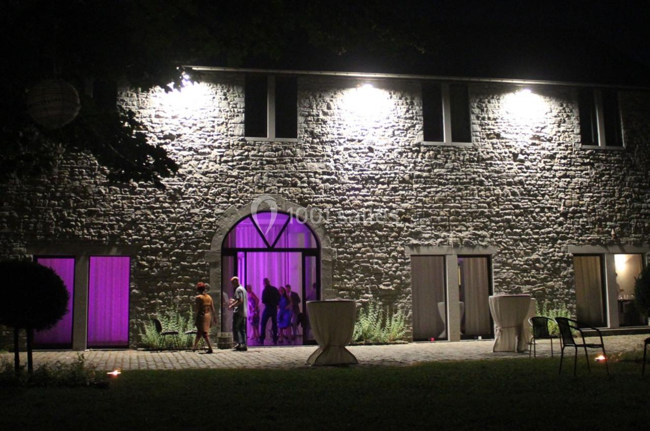 Façade en pierre d'un bâtiment éclairé de nuit, avec des fenêtres illuminées en violet et des invités entrant.