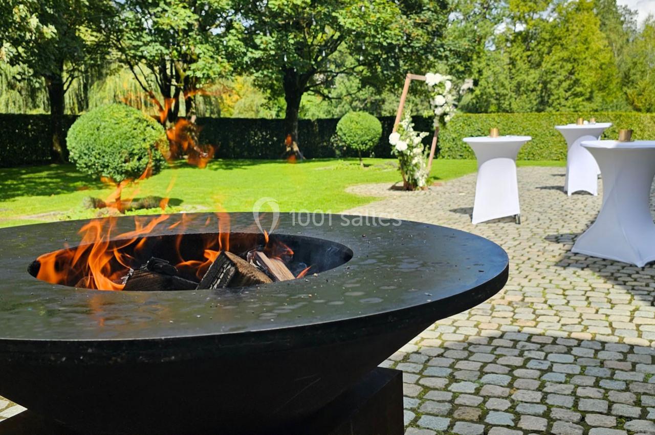 Brasero allumé sur une terrasse pavée, entouré de tables hautes et d'un jardin verdoyant en arrière-plan.