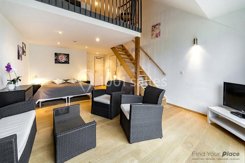 Espace de vie lumineux avec lit double, coin salon en rotin, escalier en bois et télévision sur meuble blanc.