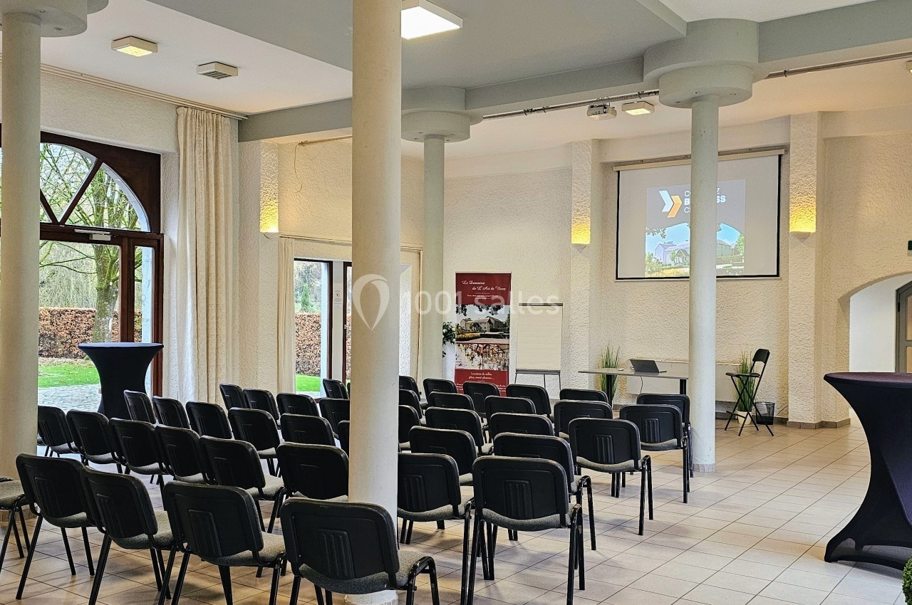 Salle lumineuse aménagée pour une présentation, avec des rangées de chaises, un écran de projection et des tables hautes.