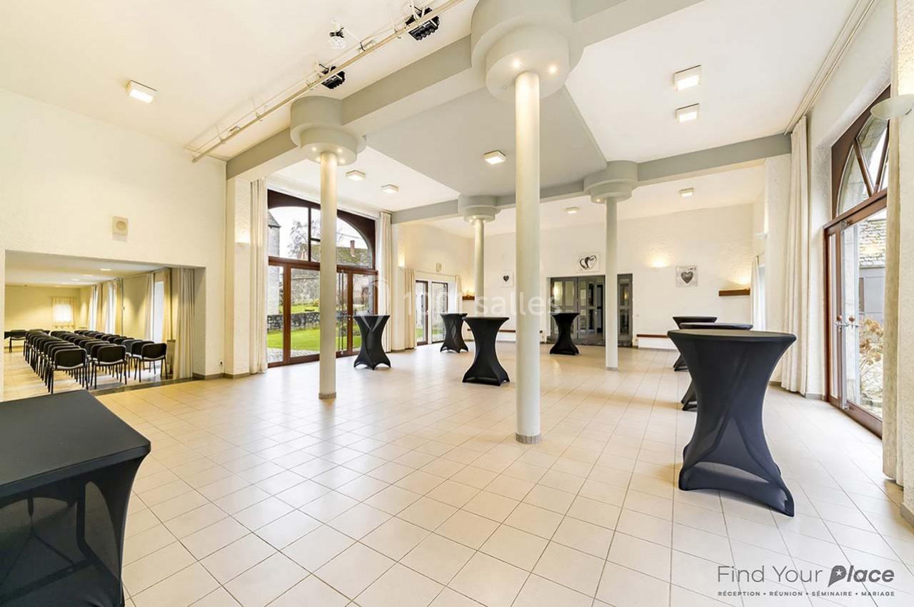 Salle lumineuse avec grandes baies vitrées, tables hautes noires, carrelage clair et vue sur un espace extérieur verdoyant.