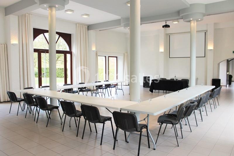 Salle lumineuse avec de grandes fenêtres, tables disposées en U, chaises noires et écran de projection en fond.