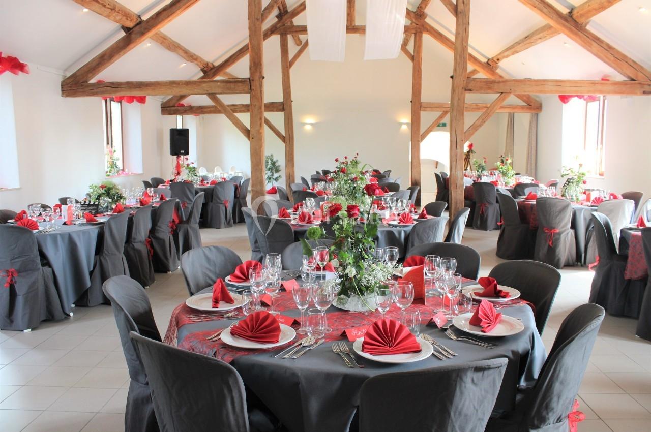 Salle de réception décorée avec des tables rondes, nappes grises, serviettes rouges pliées et arrangements floraux.