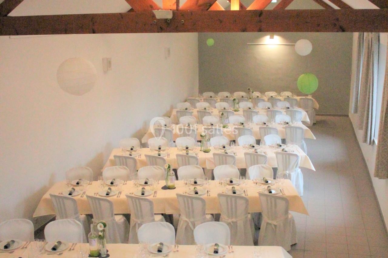 Salle de réception avec tables alignées, nappes blanches, chaises houssées et poutres apparentes au plafond.