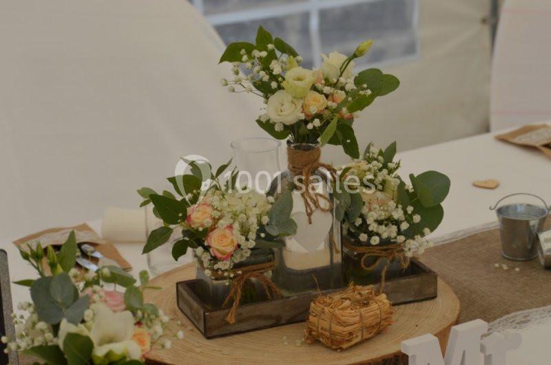 Arrangement floral avec roses, gypsophiles et feuillage dans des vases en verre sur un plateau en bois rustique.