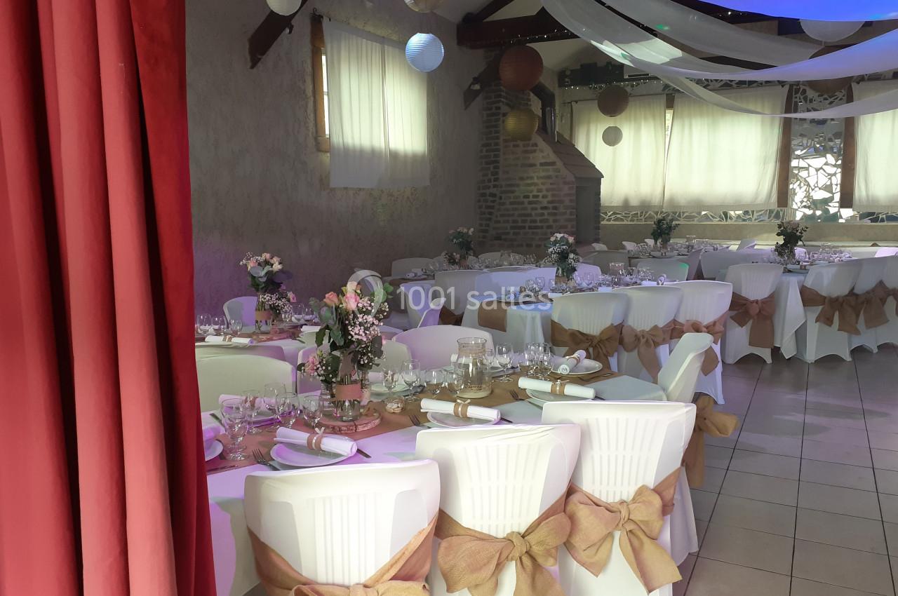 Salle décorée pour un événement, avec tables dressées, chaises ornées de nœuds en toile de jute et éclairage doux.