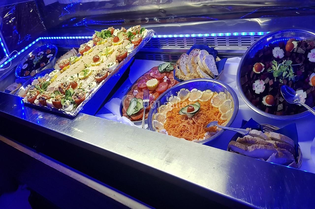 Buffet froid avec plats variés : salades, légumes, charcuterie, pain et garnitures, présenté sous éclairage bleu.