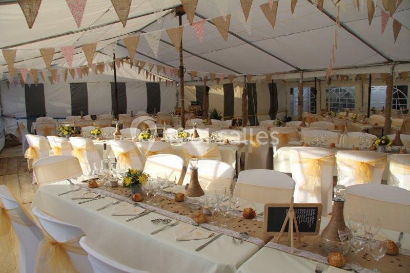 Salle de réception sous chapiteau décorée avec des guirlandes, des tables dressées et des chaises ornées de rubans jaunes.