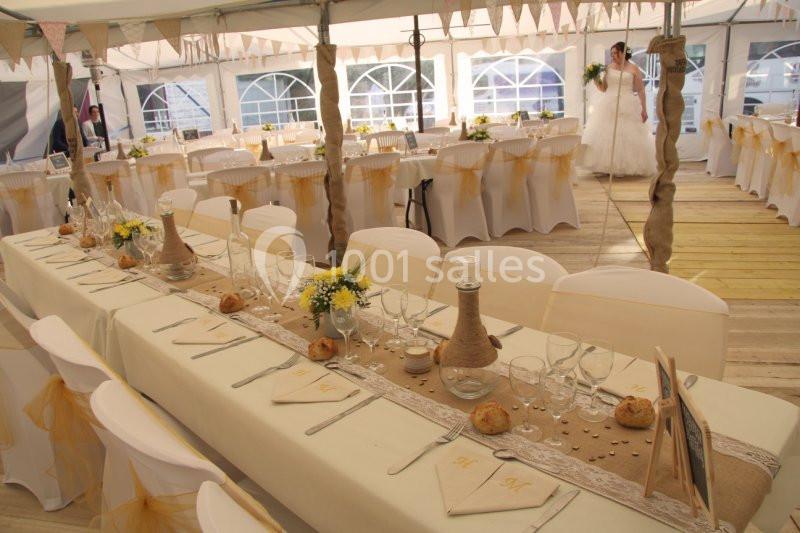 Salle de réception décorée pour un mariage, avec longues tables dressées, nappes blanches et détails champêtres.