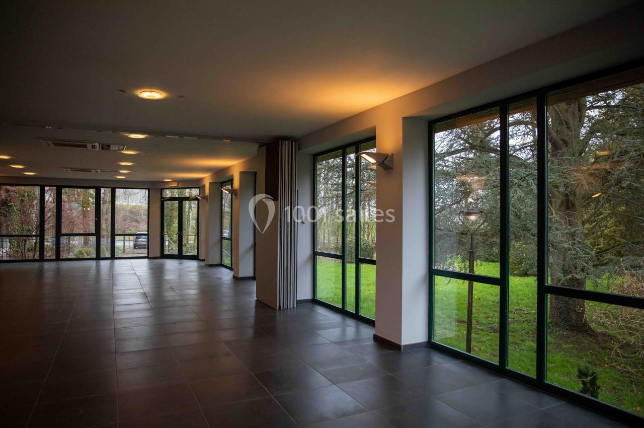 Salle vide avec de grandes baies vitrées donnant sur un espace vert, éclairée par une lumière douce.