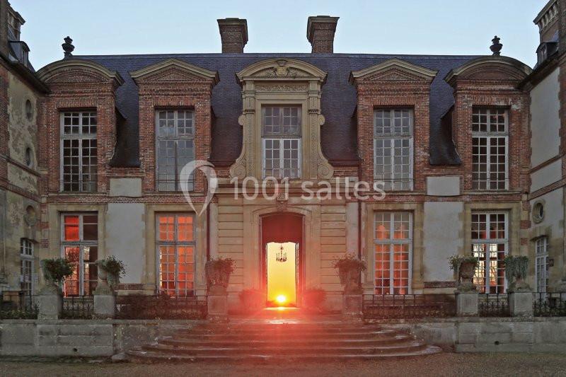 Façade d'un château en pierre et briques, éclairée par un coucher de soleil visible à travers la porte centrale.