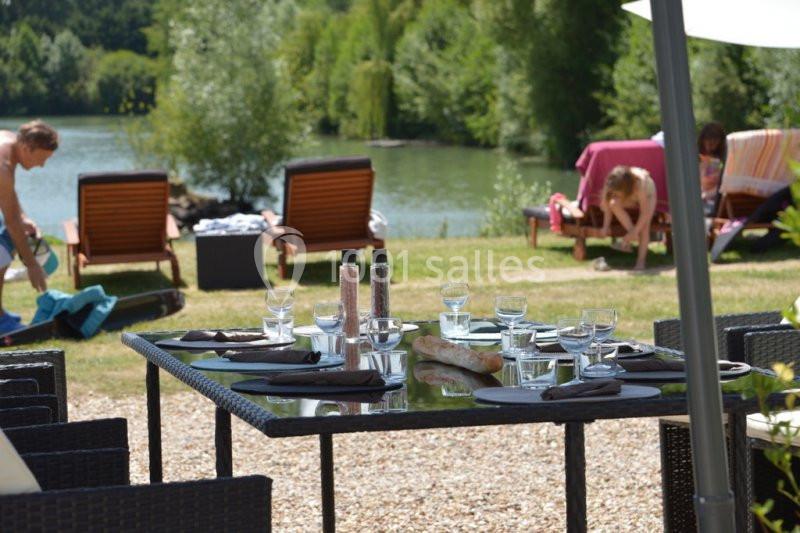 Table dressée avec vaisselle et serviettes noires en terrasse, vue sur des transats près d'un lac entouré de verdure.