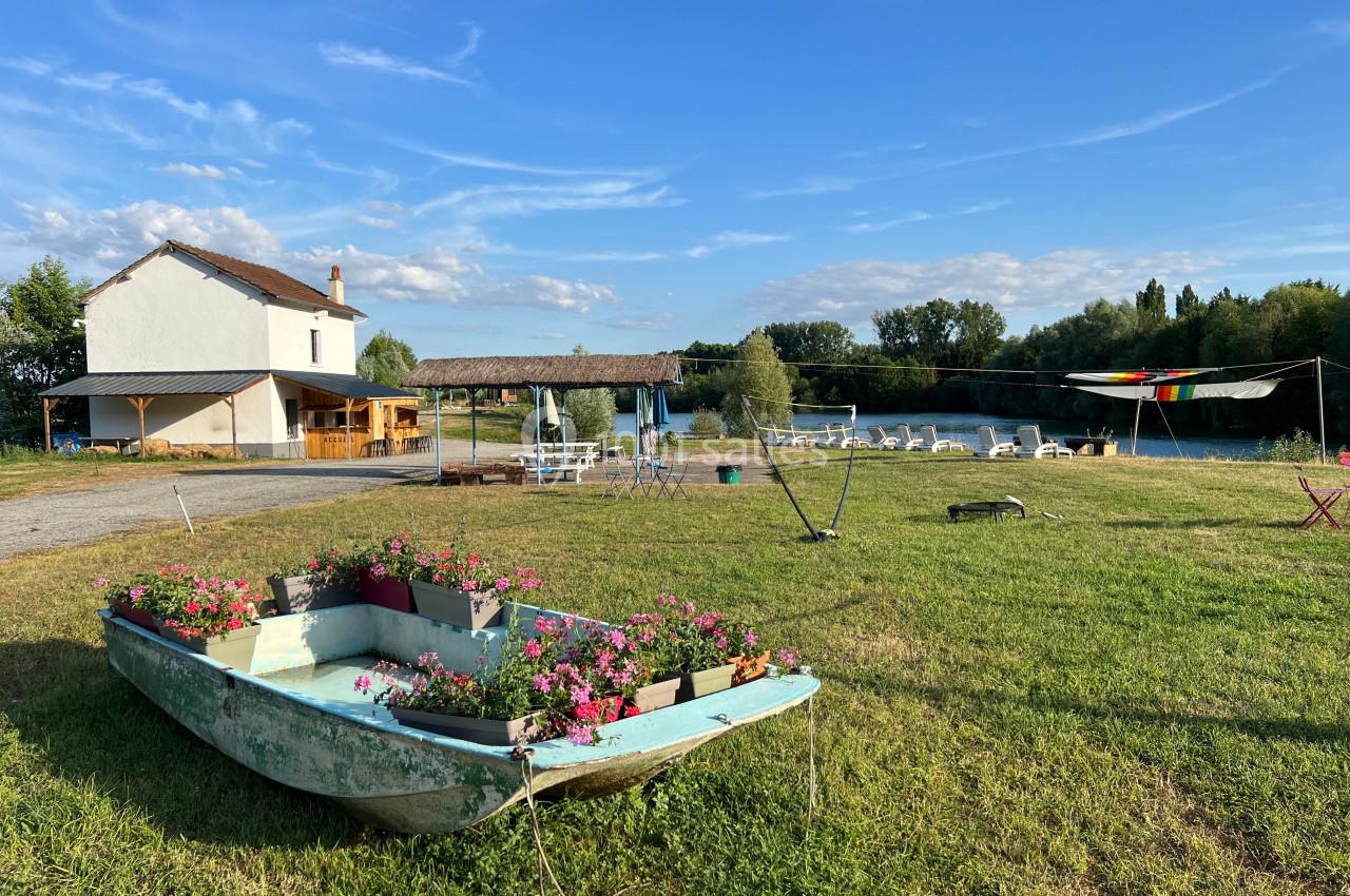 Une barque fleurie sur une pelouse, avec une maison, des tables et un cours d'eau en arrière-plan sous un ciel bleu.