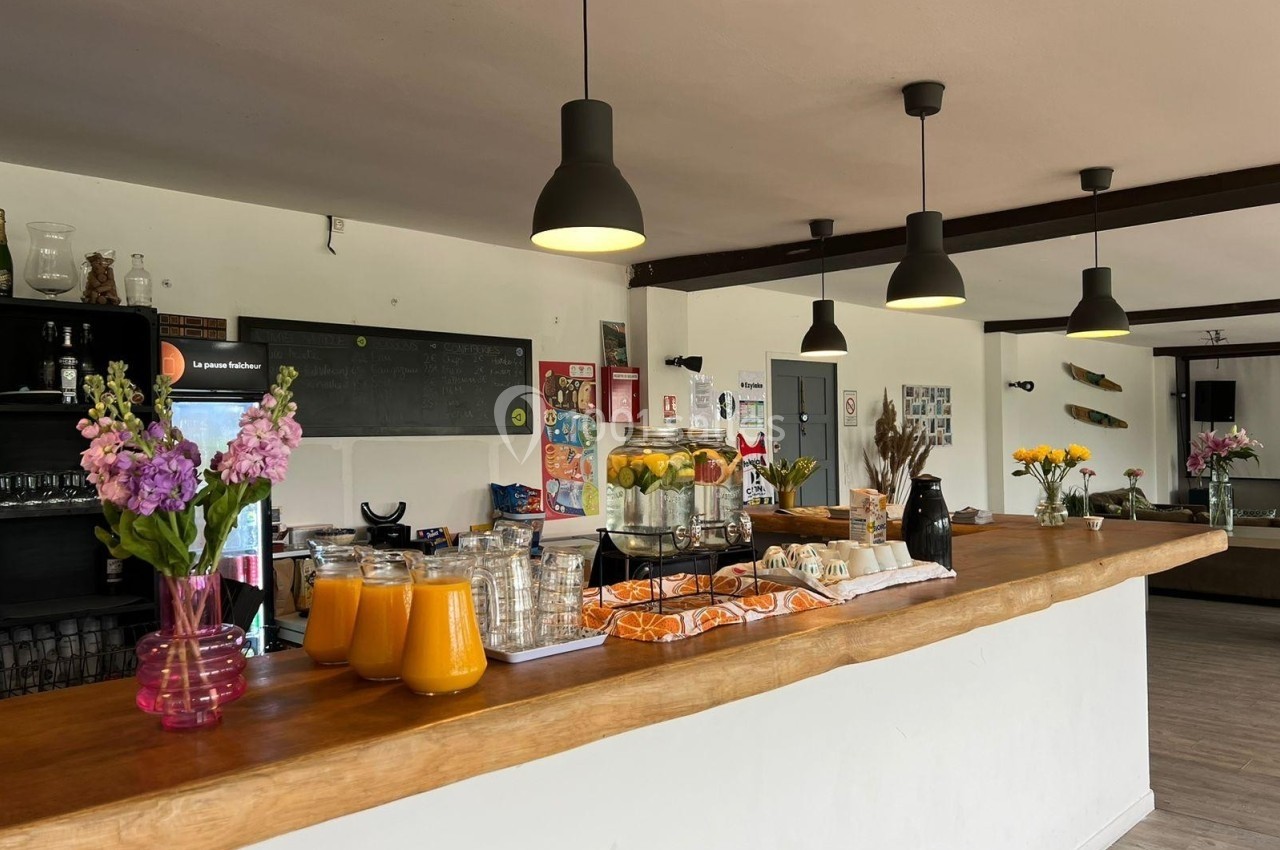 Comptoir en bois avec carafes de jus, viennoiseries et fleurs, éclairé par des suspensions noires dans un espace lumineux.