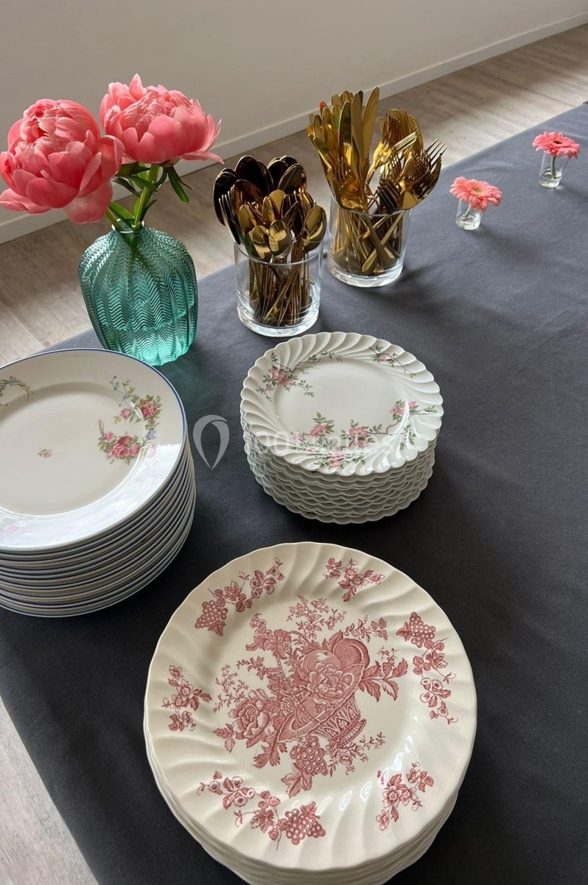 Vaisselle en porcelaine décorée, couverts dorés et vase avec des fleurs roses disposés sur une nappe noire.