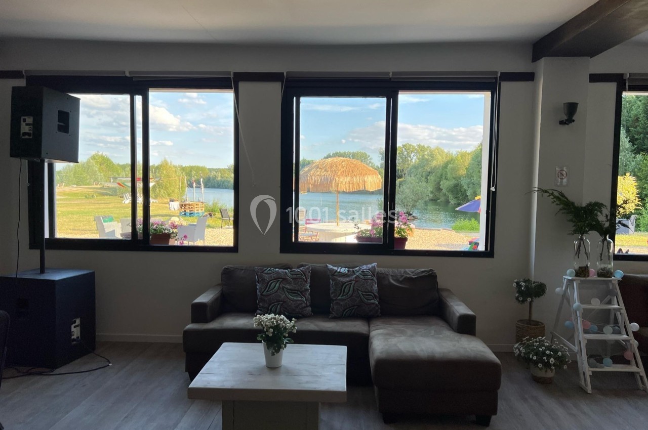 Canapé et table basse dans une pièce lumineuse avec vue sur un lac, des parasols et un paysage verdoyant à travers les…