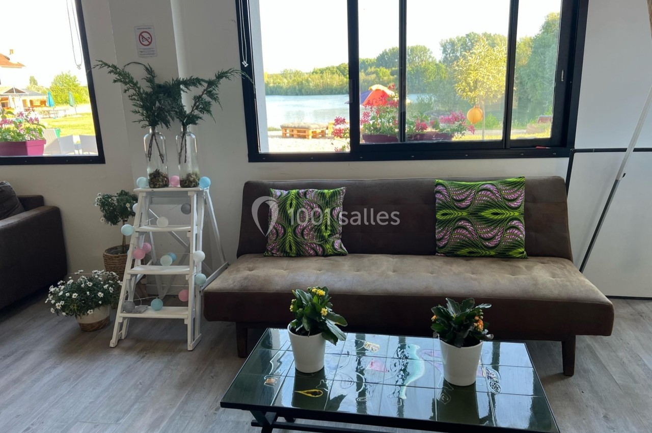 Salon lumineux avec canapé brun, coussins colorés, table basse avec plantes et vue sur un lac à travers de grandes fenêtres.